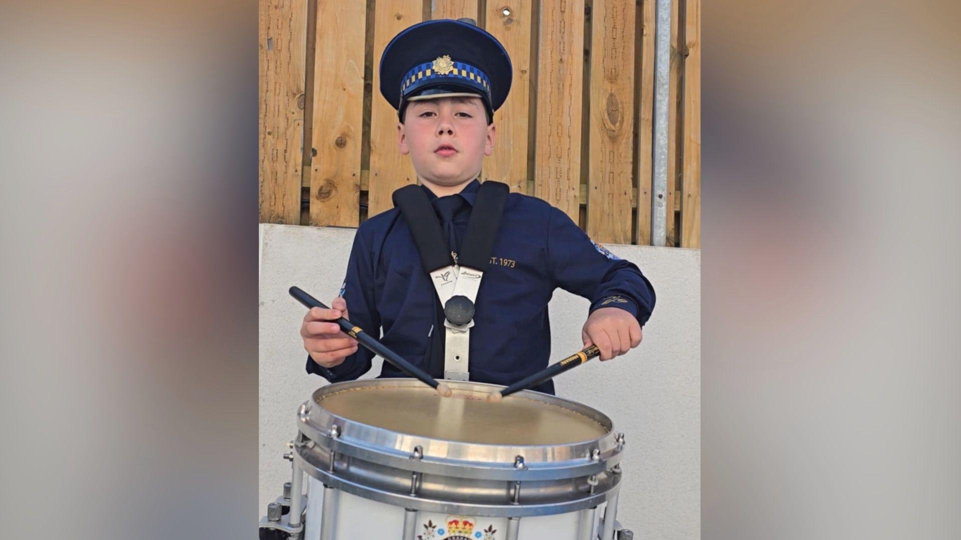 A boy wearing a navy bandsman's uniform and a hat holds a drum, held around his neck and in his hands are two drum sticks. He stares at the camera with a neutral expression on his face.