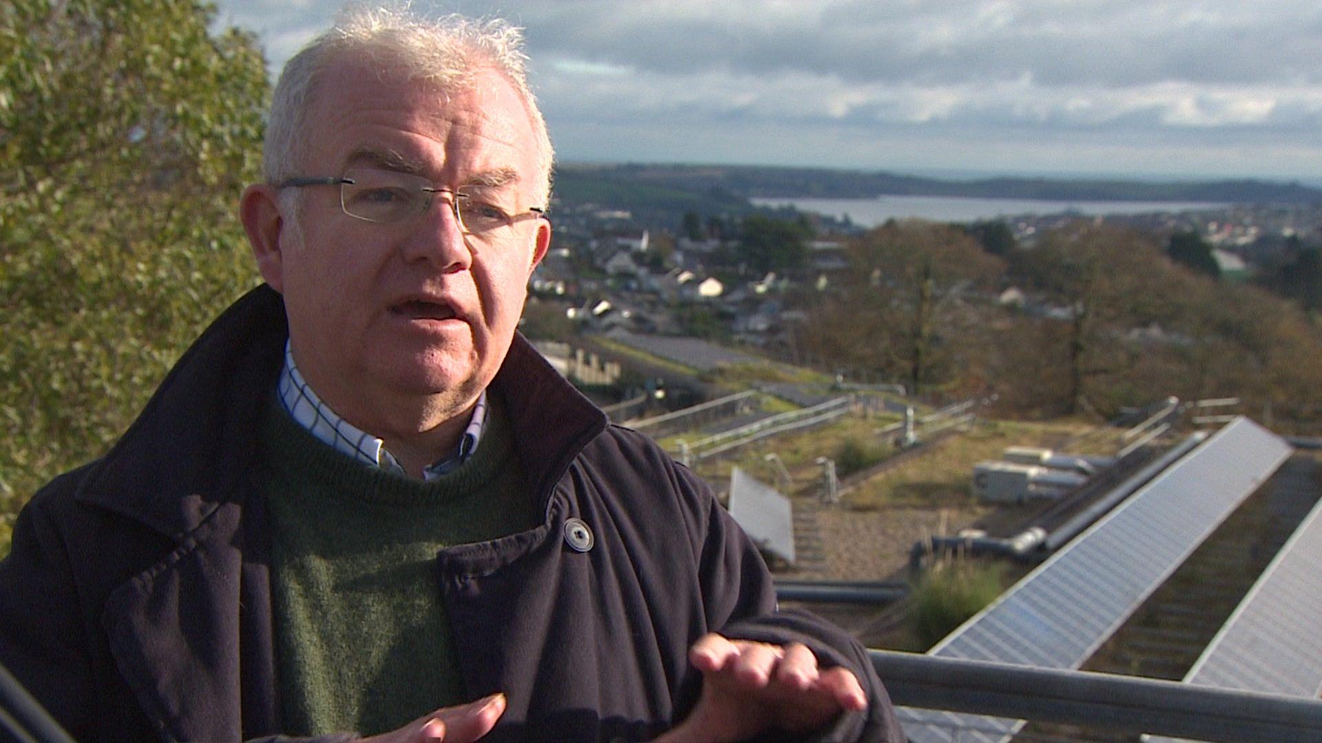 A man with grey hair and glasses wearing a green jumper and black coat standing in front of some solar panels on a building roof behind him 