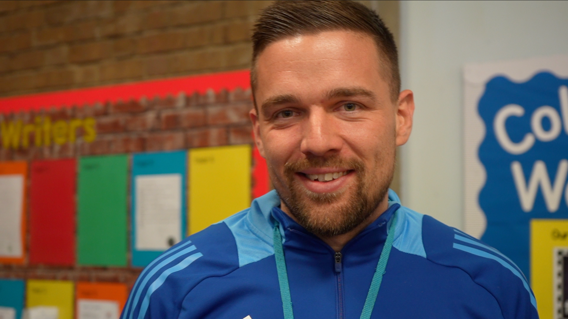 Ben Andrews smiles at the camera while standing in front of several school notice boards. He has short dark hair with a dark beard. He is wearing a blue jacket.