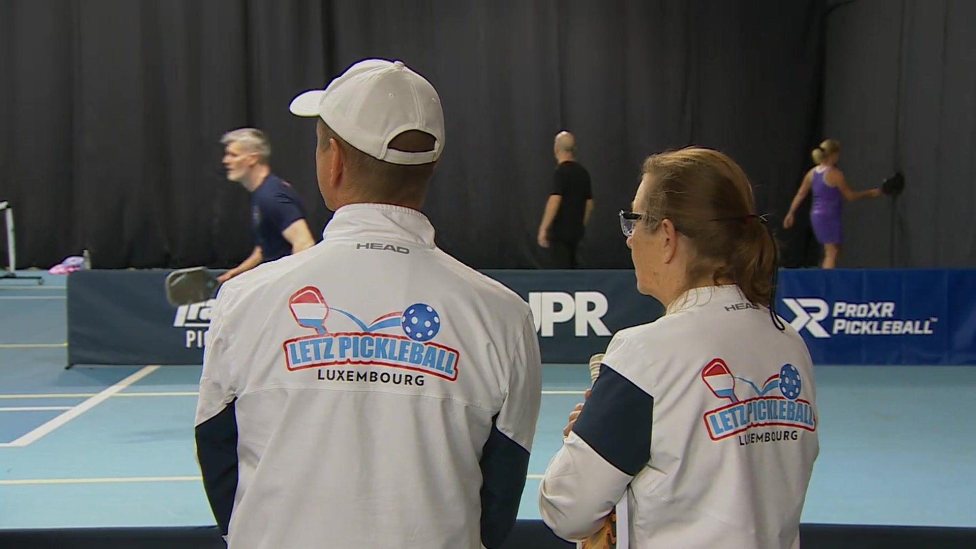 Two pickleball players from Luxembourg are watching other players on the court. They have their backs to the camera.