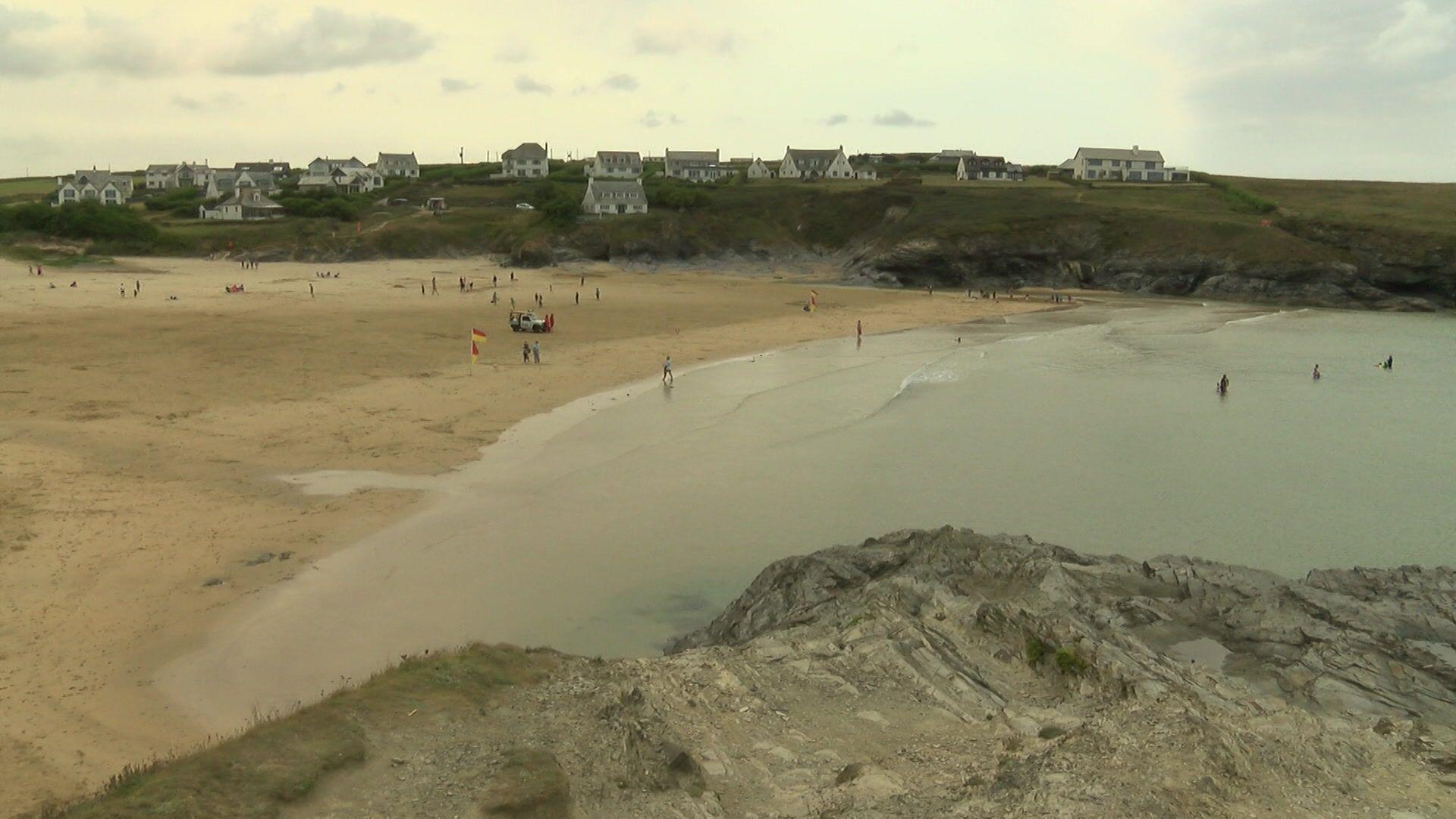 A picture overlooking a sandy beach which has people on it. The sea is blue and at the forefront of the image is a rocky cliff. In the distance is green grass and houses dotted along it. In the distance, at the centre of the beach, there is a red and yellow flag.