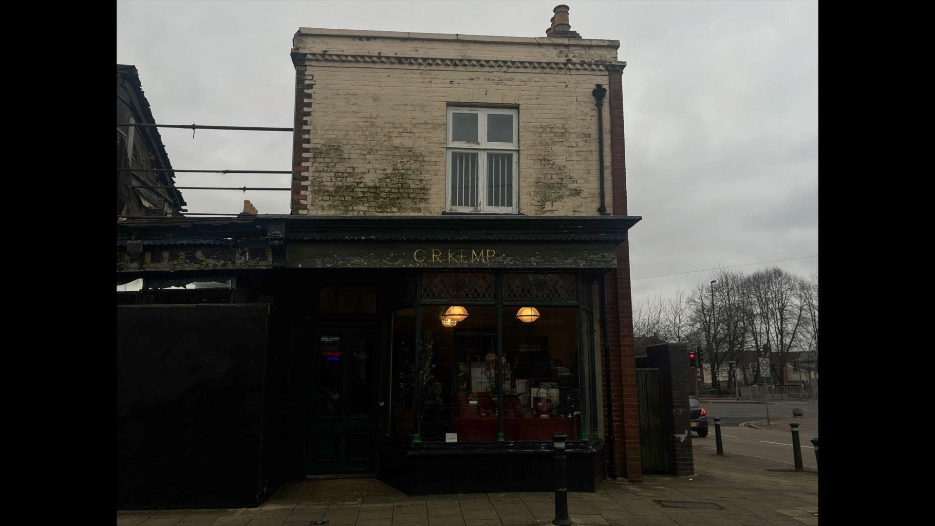 A two-storey building. The lower flood has a black shop front with bars on the windows. The upper flood has one window with white bars. It is an end of terrace property. The building next door has been partially demolished with bracing bars in place.
