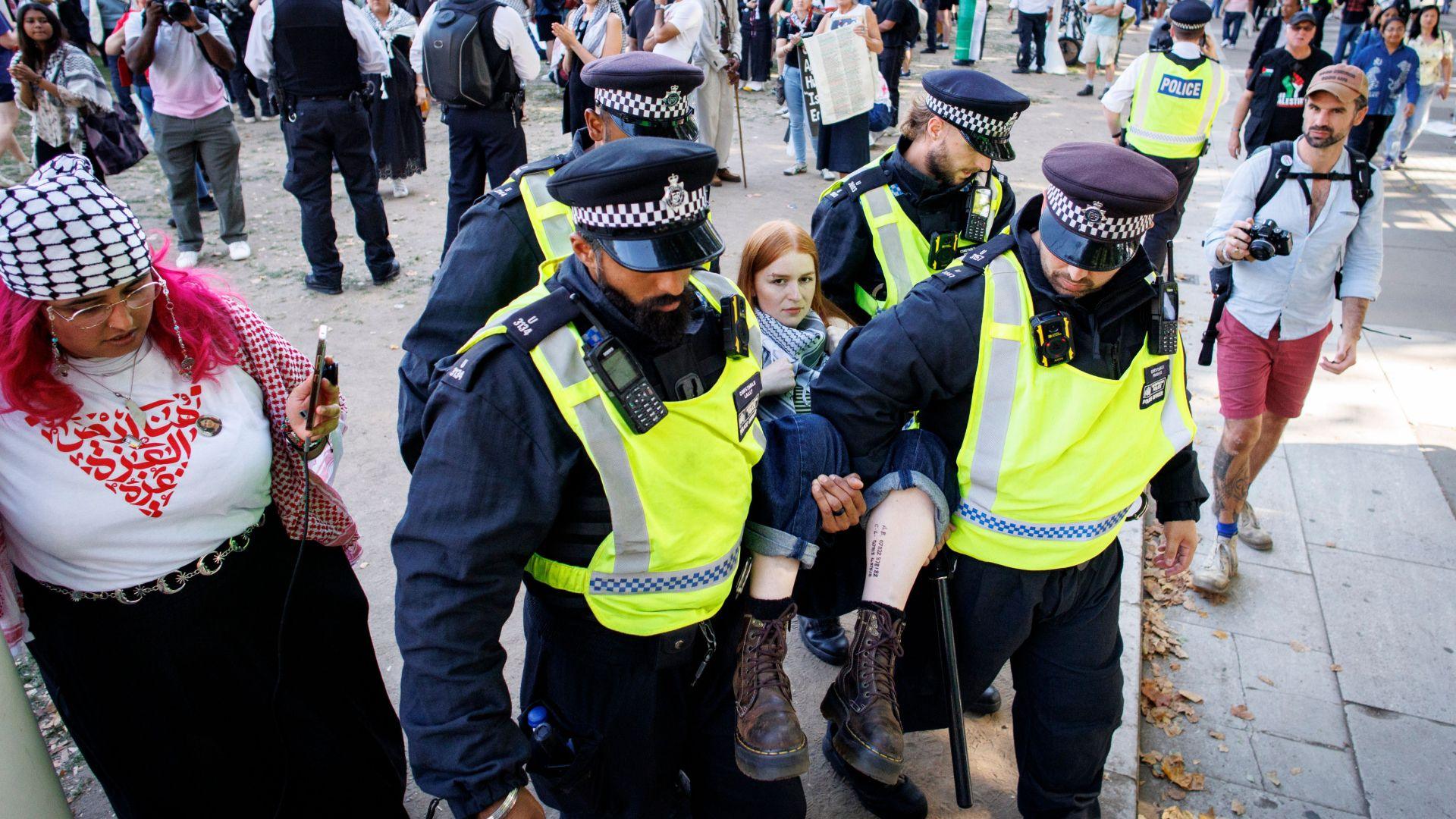 A young woman with red hair is carried by four male police officers, with each holding one of her four limbs, as a crowd of protesters is seen behind them.