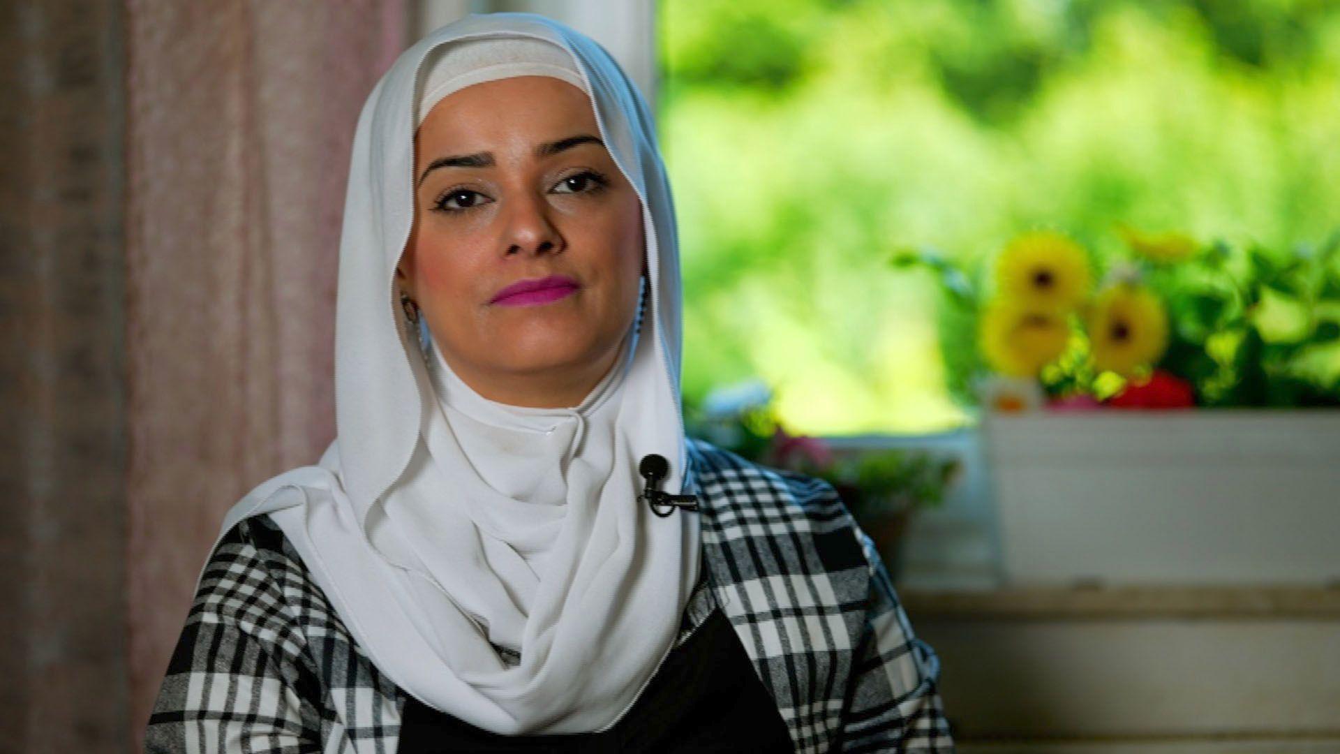 Headshot of a woman wearing a black and white checked top with a white headscarf. She is wearing pink lipstick. There is a pot with yellow flowers in the background.