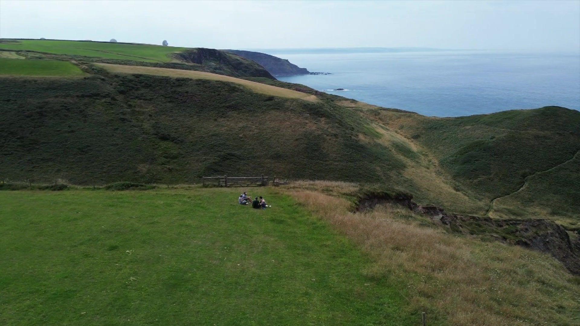 Landscape picture showing some people sitting on a vast expanse of grass next to cliffs. The sea can be seen to the right.