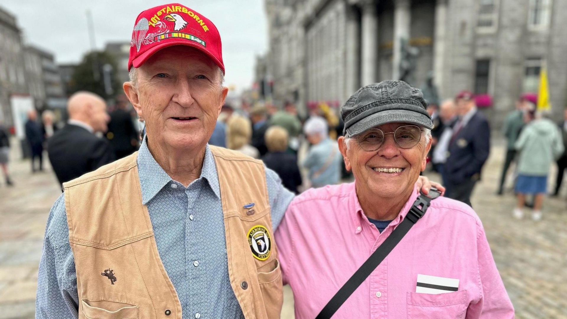 Two elderly men, standing with their arm round each others shoulders. One is wearing a red cap with military details on it and a beige vest. The other is wearing a pink shirt and grey cap.