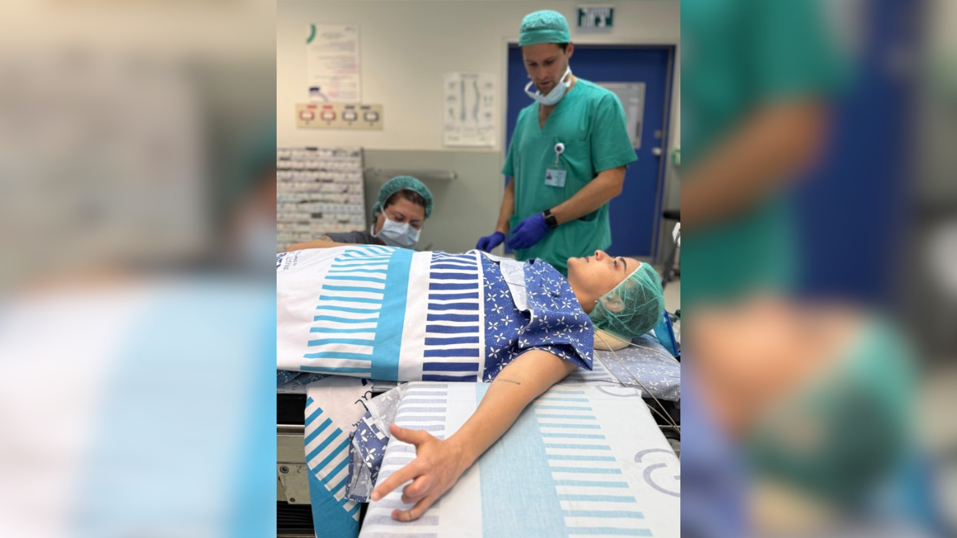 Emily Damari lays on an operating table wrapped in hospital blankets, with two health workers dressed in green behind her.