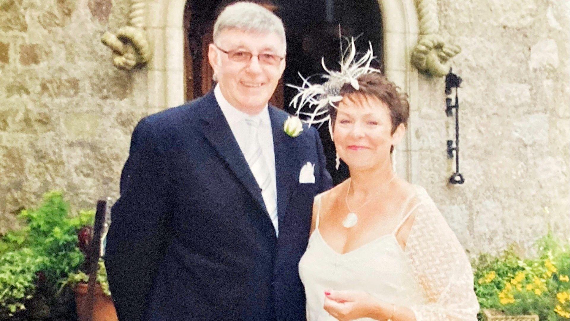 Man - Stan Shepherd - and his wife Susan Shepherd, he is dressed in a smart dark suit and she is wearing a cream dress, both smiling at the camera, at a wedding.