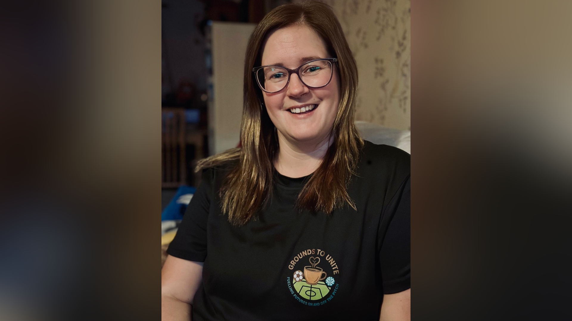 Liza smiles at the camera as she sits on a sofa. She has long brown hair, glasses and wears a black T-shirt with the Grounds to Unite logo on the chest.