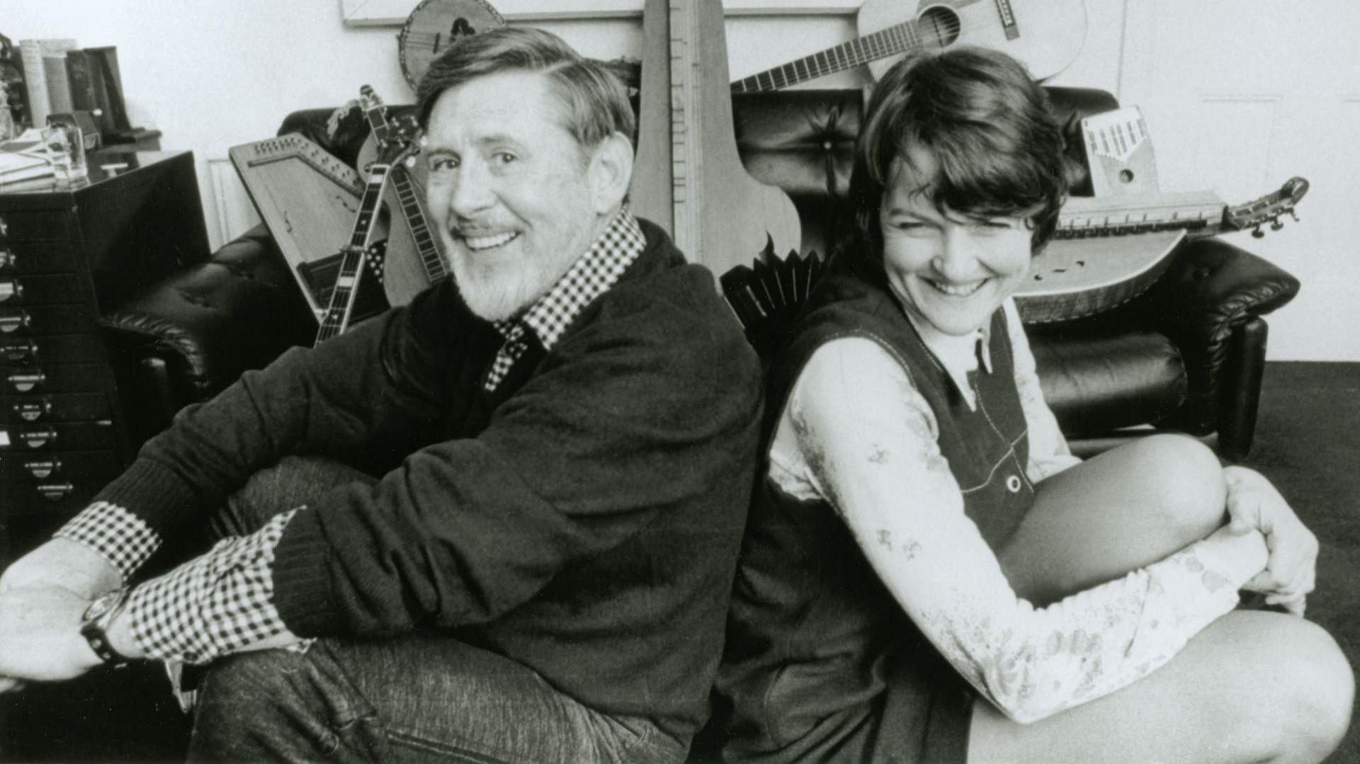 An archive Peggy Seeger and Ewan MacCall from1980. They are sitting in a room full of musical instruments. Both are smiling but only MacCall is looking at the camera.