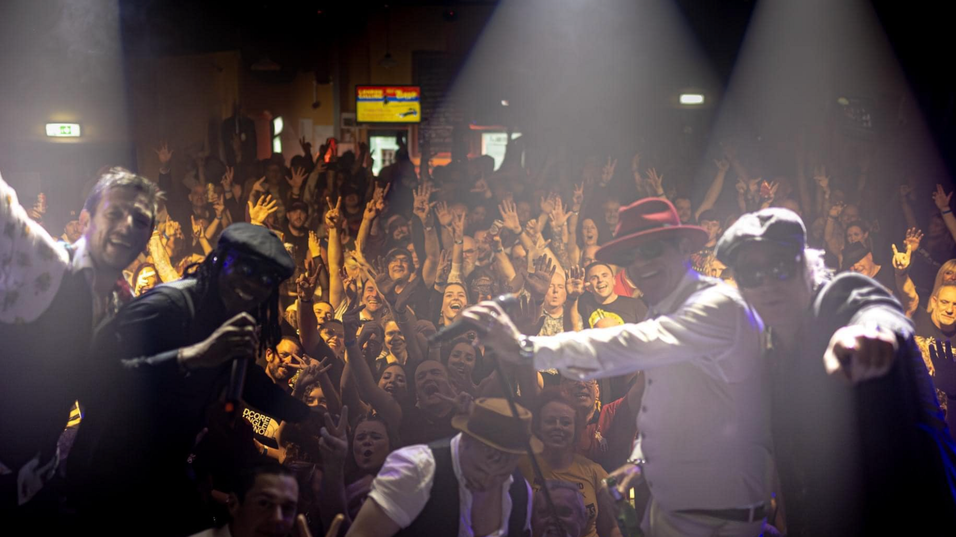 A crowd of people, lit in a yellow light all with their hands up. Everyone is posing to the camera. At the forefront of the image is four musicians, all posing for the camera.