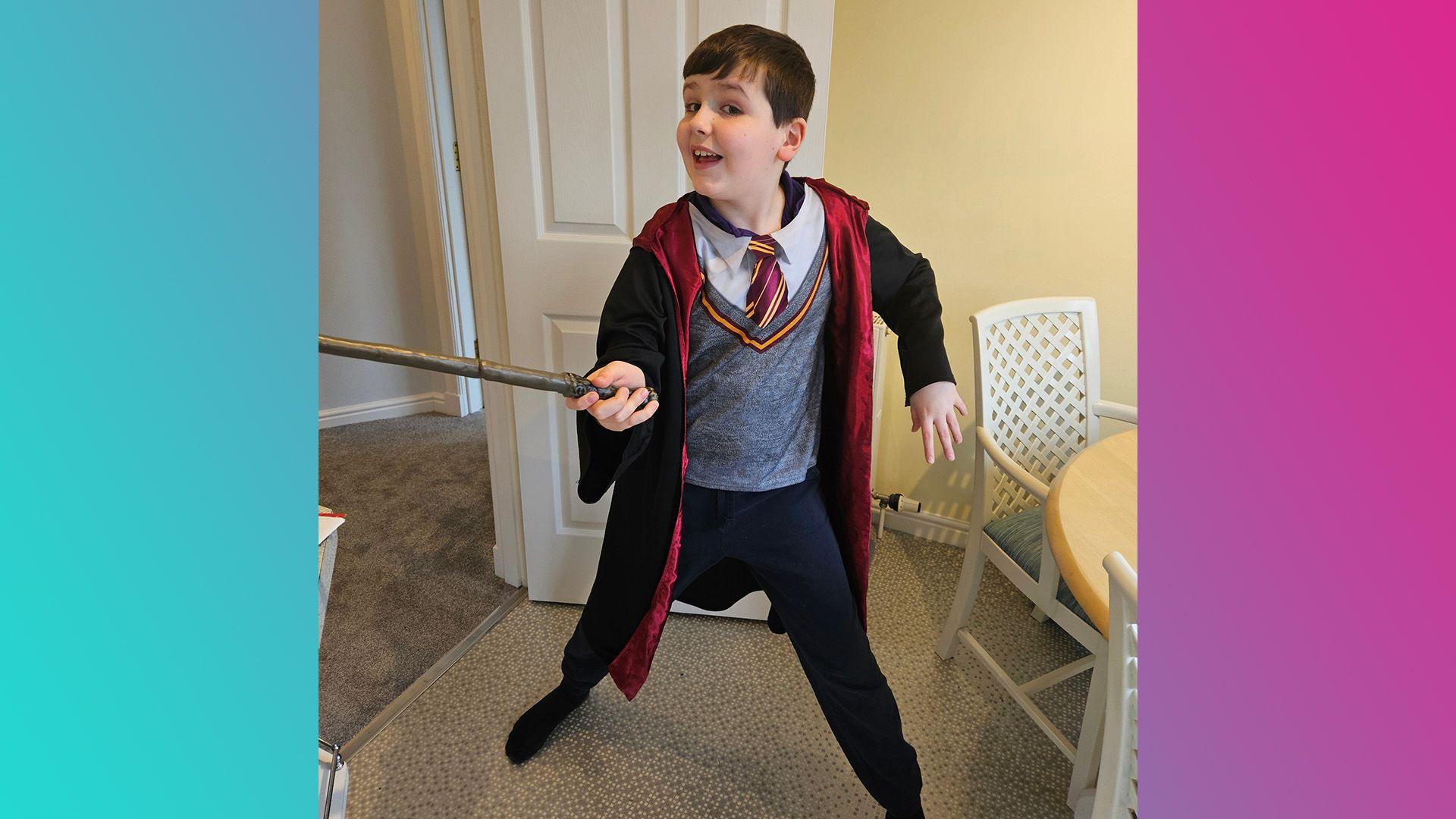 Matthew is wearing black trousers, a grey jumper and white shirt. His tie is in red and gold and he is wearing a cloak with red lining. He is holding and wand and posing to the camera as if he is casting a spell 