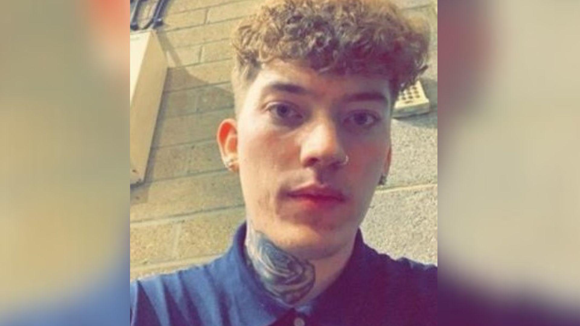 A selfie of Henry Smith, a 27-year-old man with blond hair which is short on the sides and curly on the top. He has a nose ring and a neck tattoo of a rose. He is wearing a navy shirt buttoned to the top and is sat against a wall made of large bricks.
