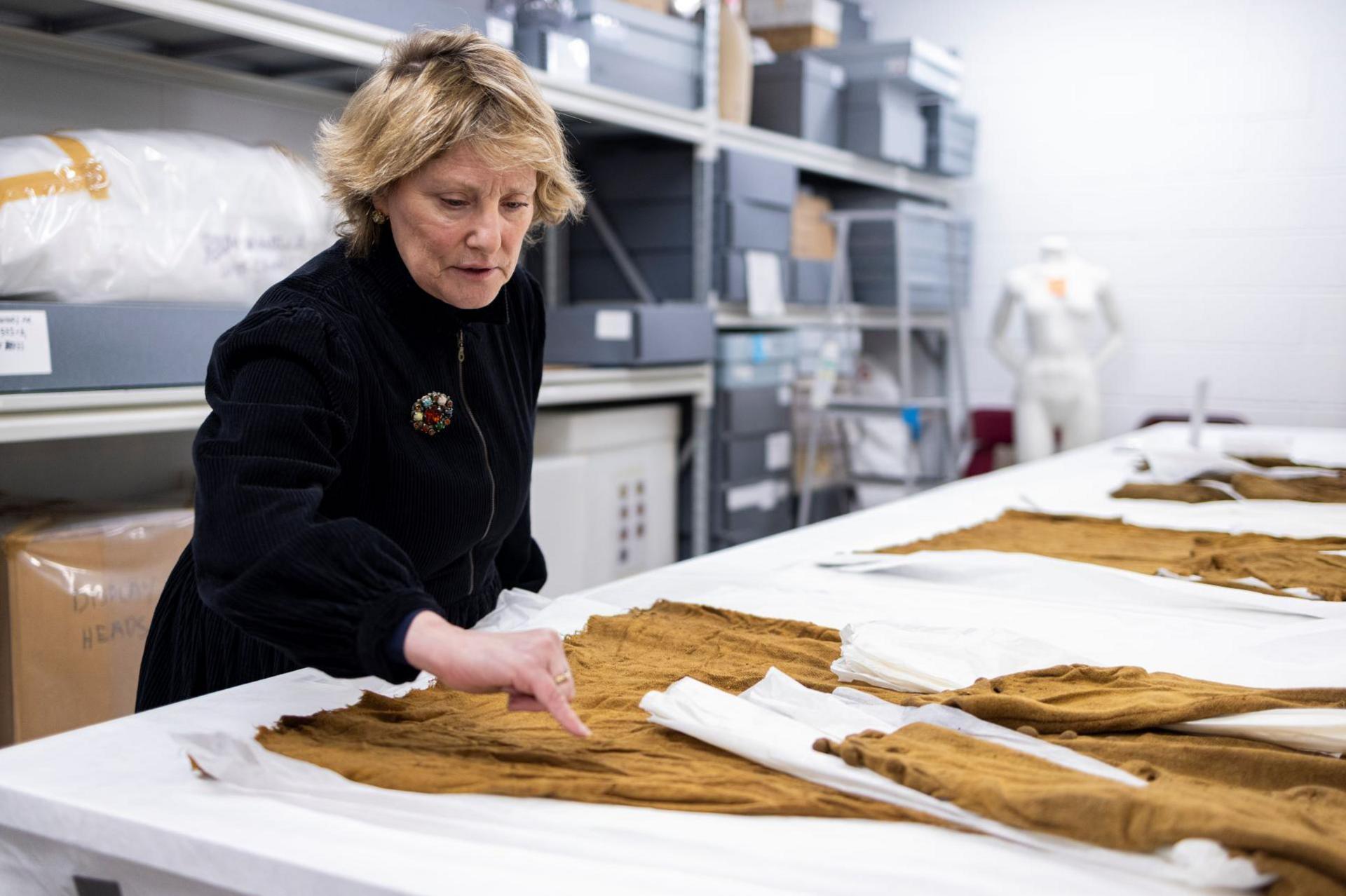 The curator, a woman who is wearing a black coat with a brooch pinned to it, points to detail on the jacket with her pinkie. The jacket and other garments are laid out on a white table.