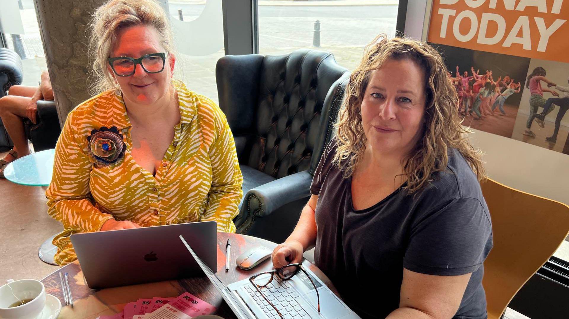 Miranda Acres (left) and Samantha Sherman from Ipswich.love in a cafe in front of laptops and looking up, the one on left wearing a yellow top and the one on the right wearing a black t-shirt