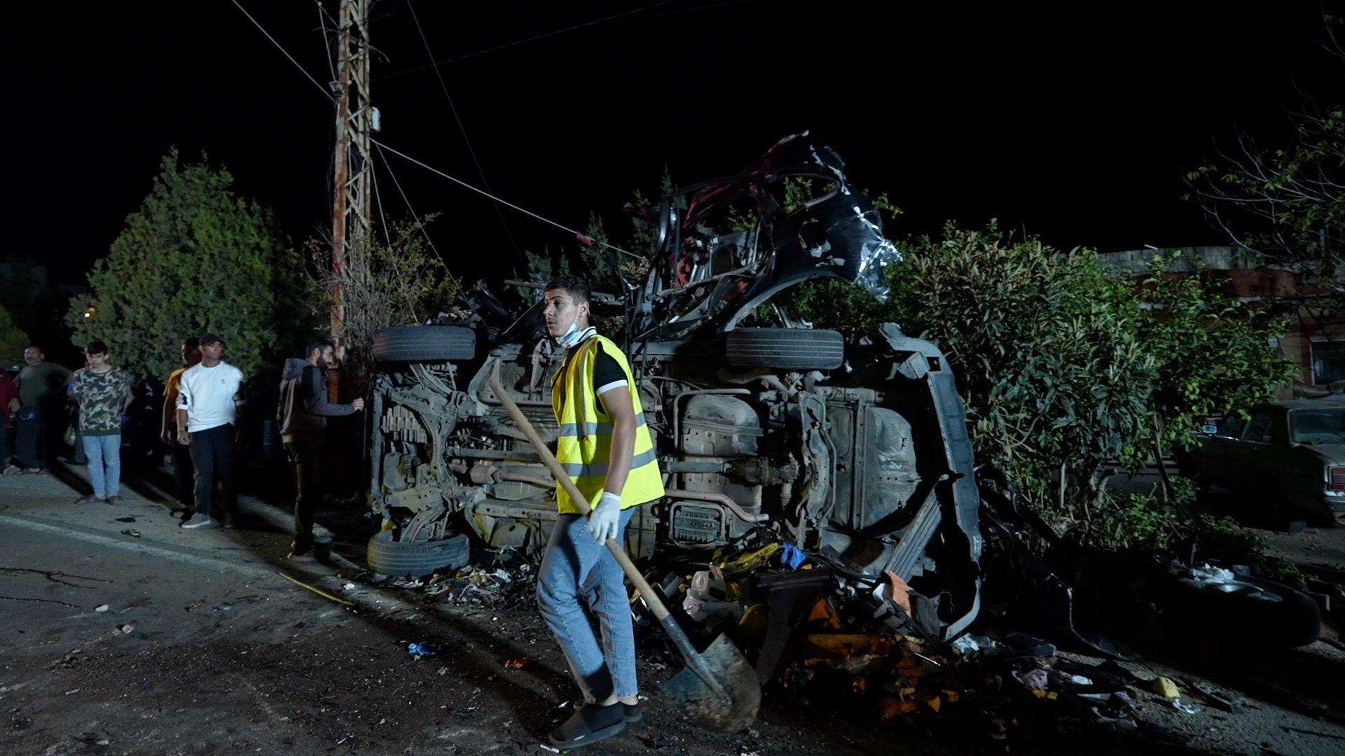 A young man in hi-vis holds a shovel in front of a car, on its side and destroyed by an apparent explosion. Other young men look on. It's dark and the car is on a street in front of a hedge.