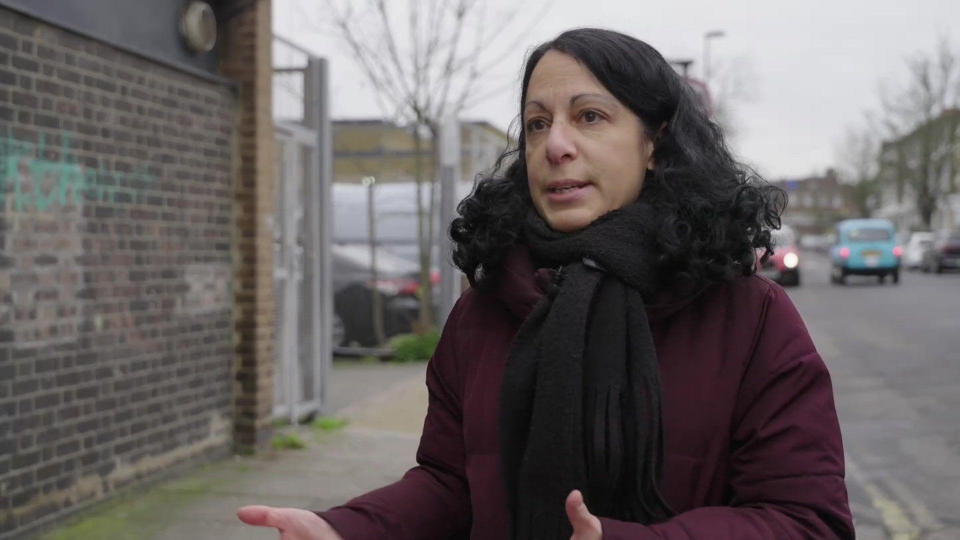 Emma Haddad standing outside on a London road. She wears a burgundy coat and black scarf. She has dark, curly hair.