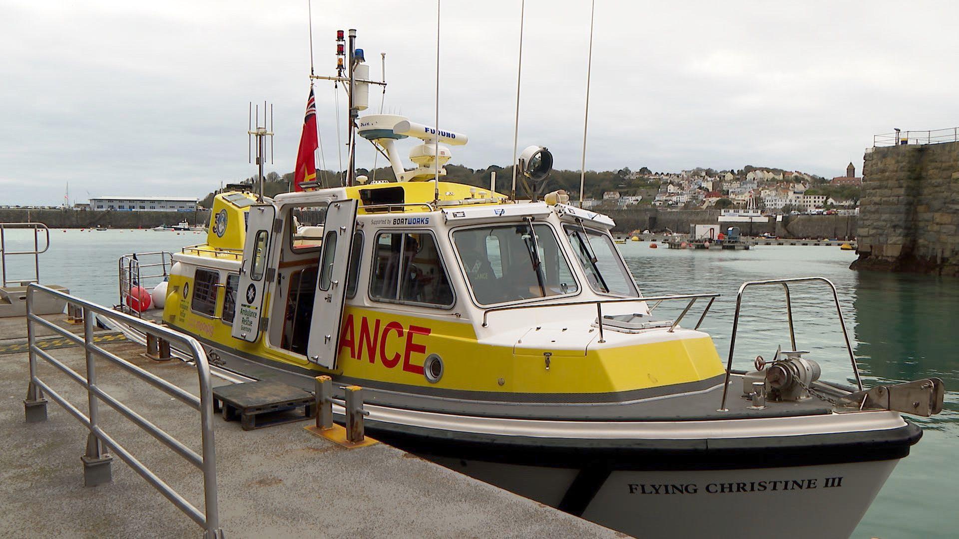 An ambulance boat. It is yellow and white. It says "Ambulance" on it. It's doors are open. There are people inside. It is docked. The water is a light blue. There are houses in the background. 