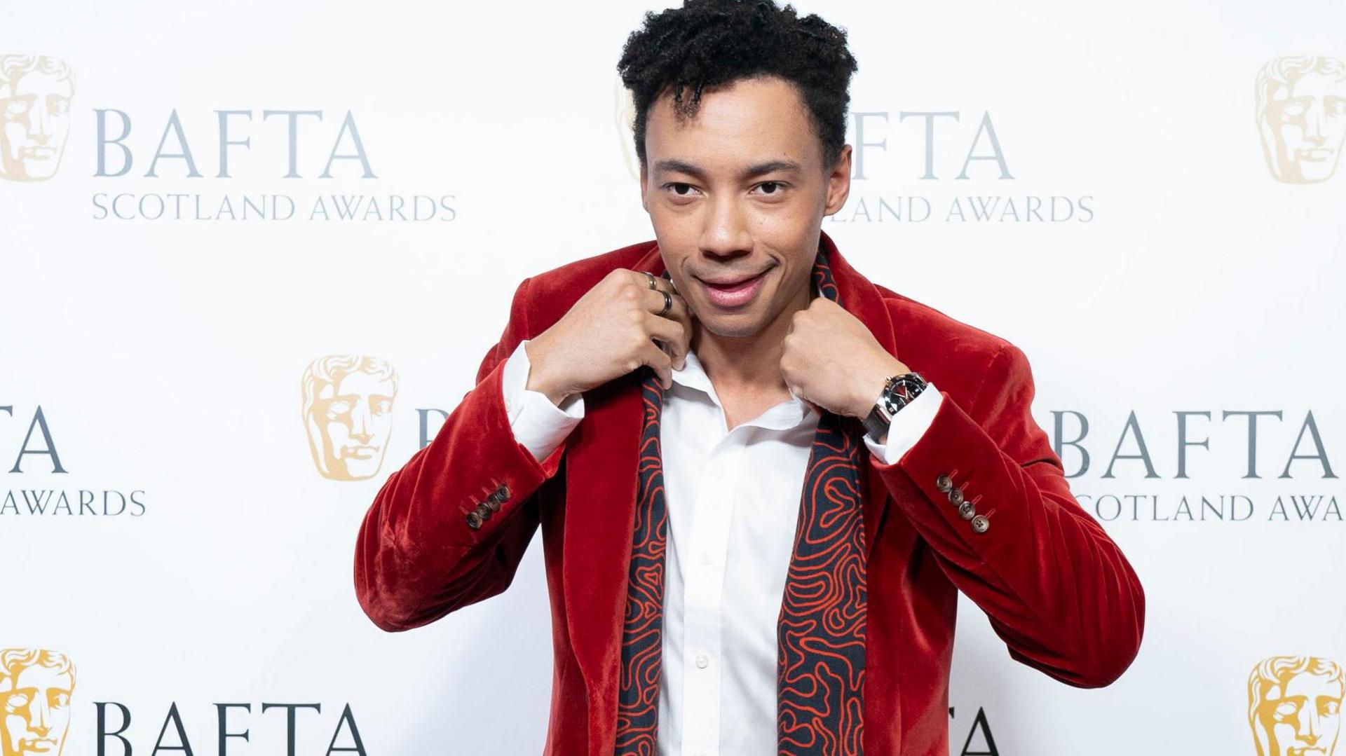 Kit Young poses at the Bafta Scotland Awards, wearing a red velvet suit and white shirt.