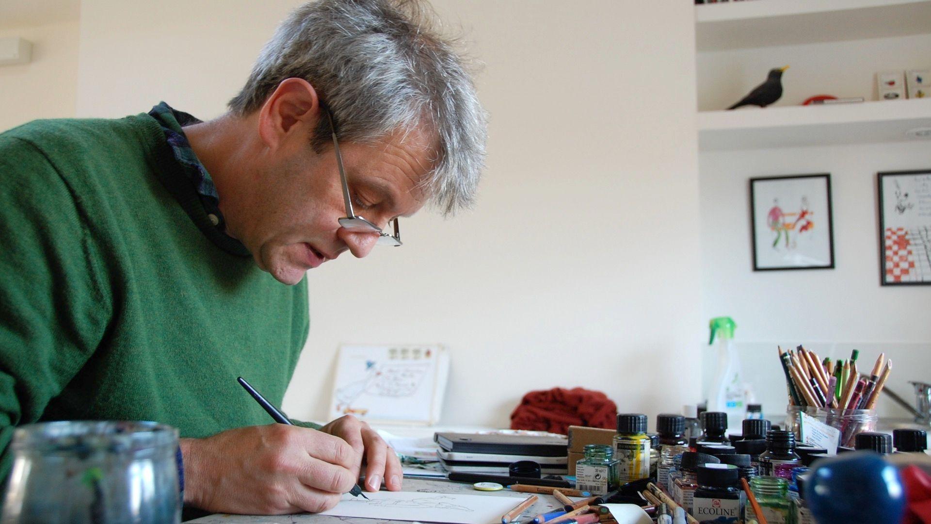 Axel Scheffler, wearing a green jumper mid-drawing, in his studio. There are paint pots and brushes on his desk