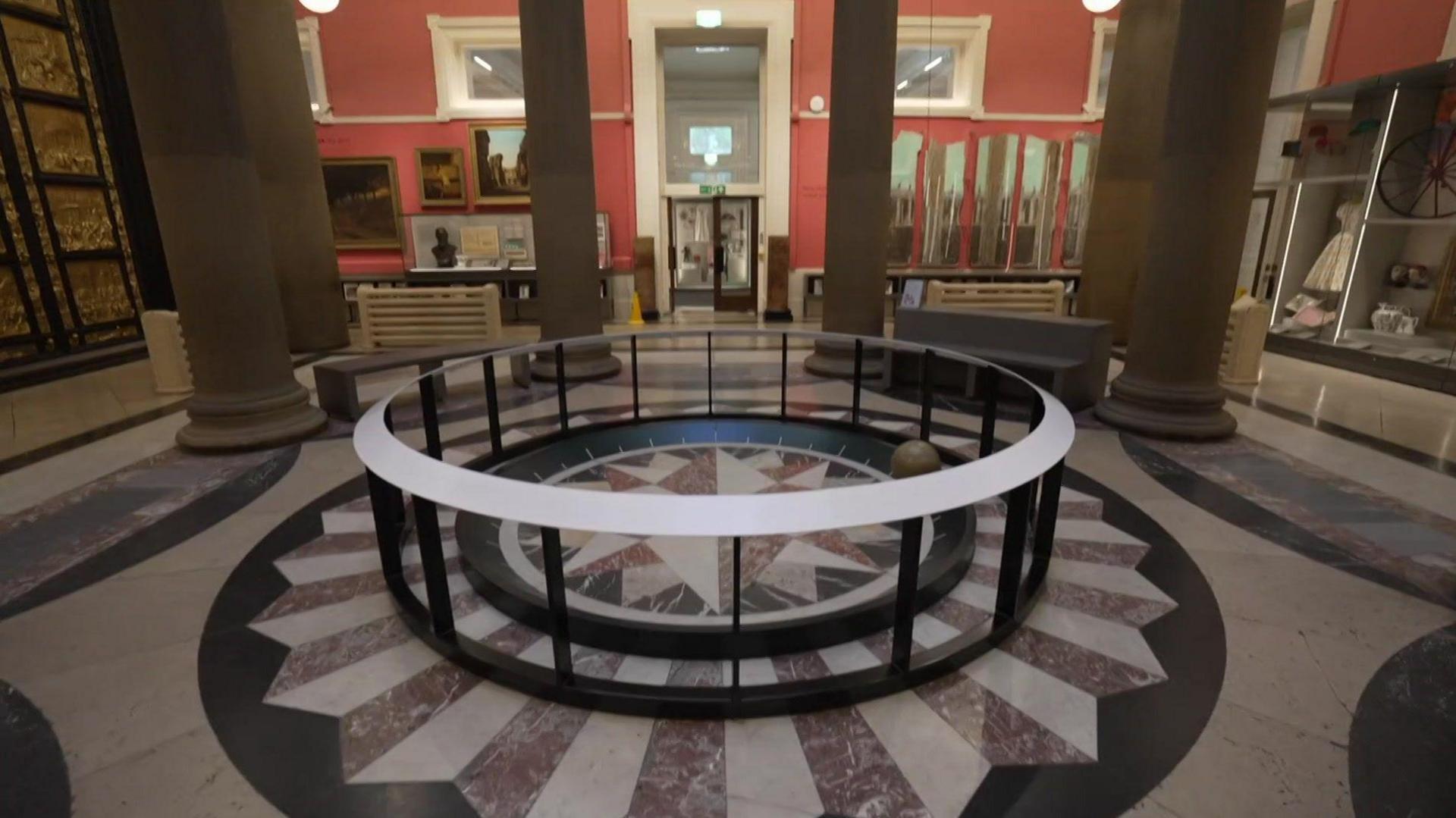 Interior of the museum with exhibits in a spacious columed hall. A large door with golden columns stands on the left and a ring surrounds the purple and white striped marble floor.