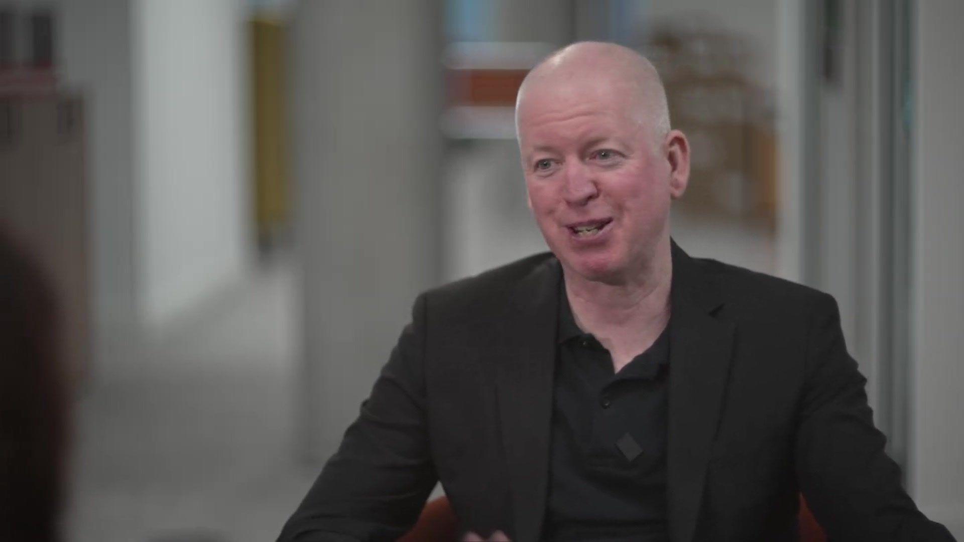 Michael Keating in a black suit jacket and black polo t-shirt.