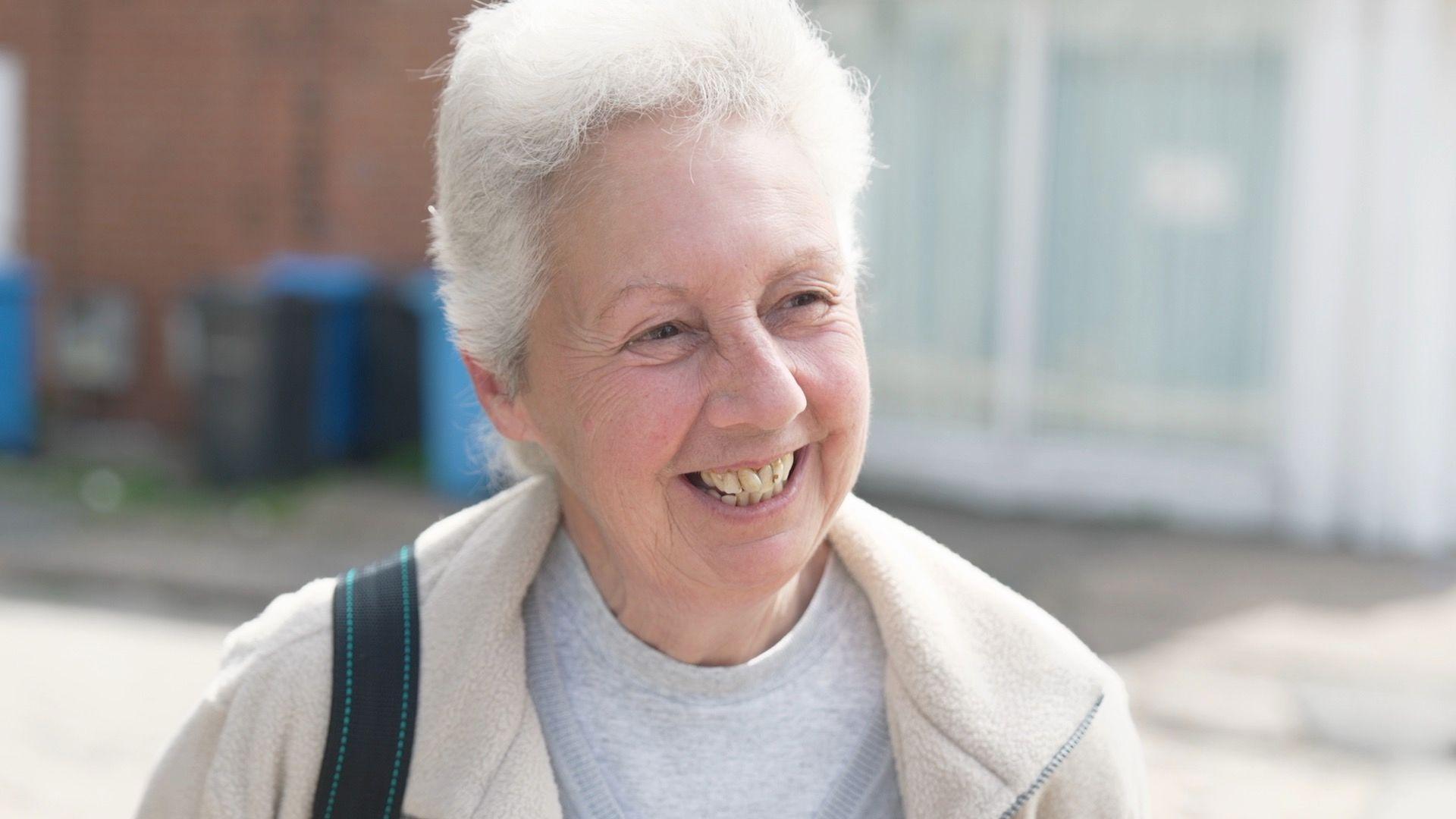A smiling woman with grey hair stands in front of bins, wearing a grey t-shirt and cream top. She has a black satchel strap on her shoulder.