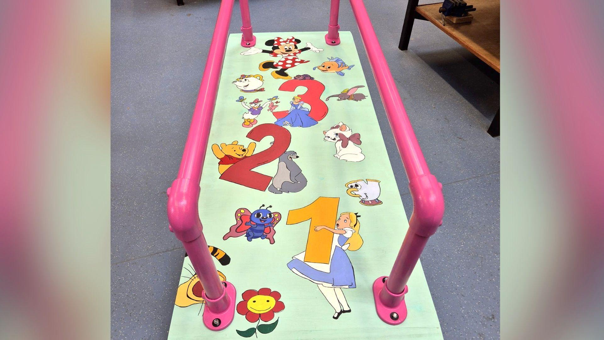 A walking mat with pink parallel metal bars, used for young children with mobility issues to practice walking. The mat is decorated with cartoon characters such as Alice in Wonderland, Minnie Mouse and Winnie the Pooh. 