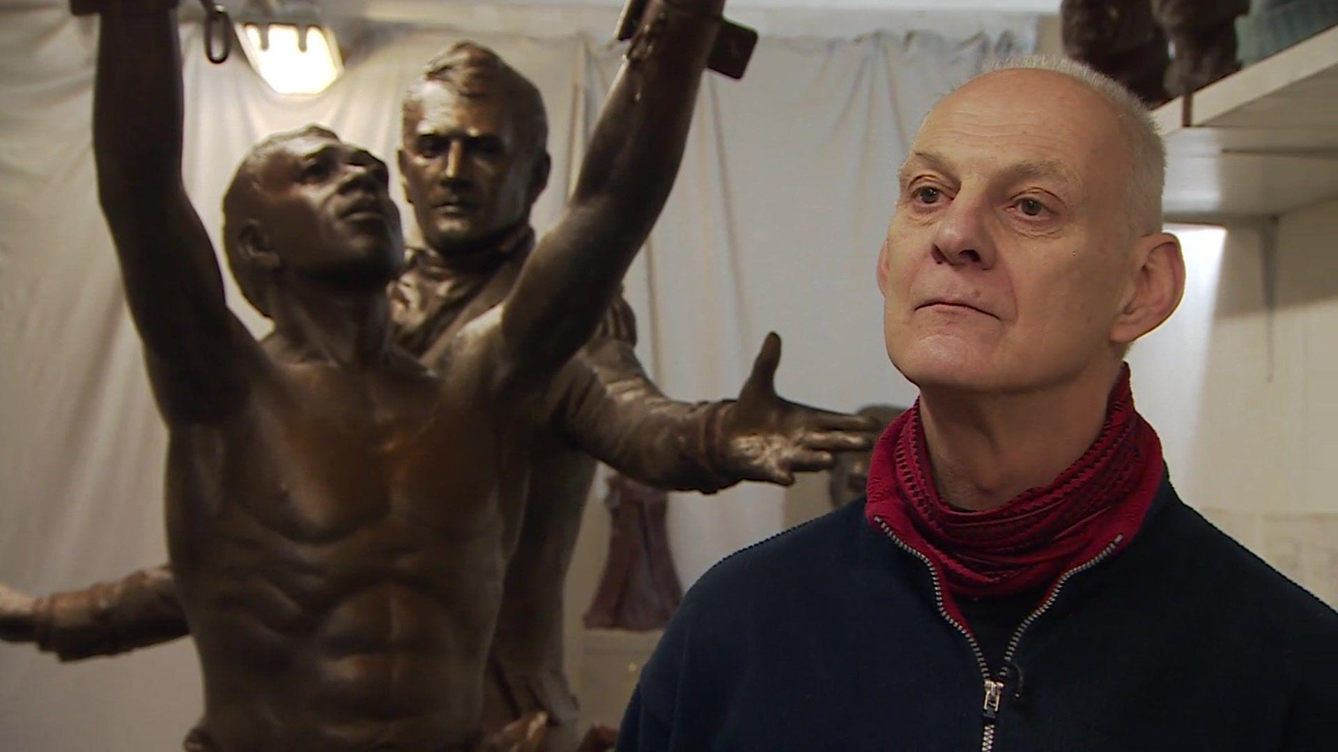 Vincent Gray stands in front of a bronze sculpture that is in front of a backdrop of white sheets.