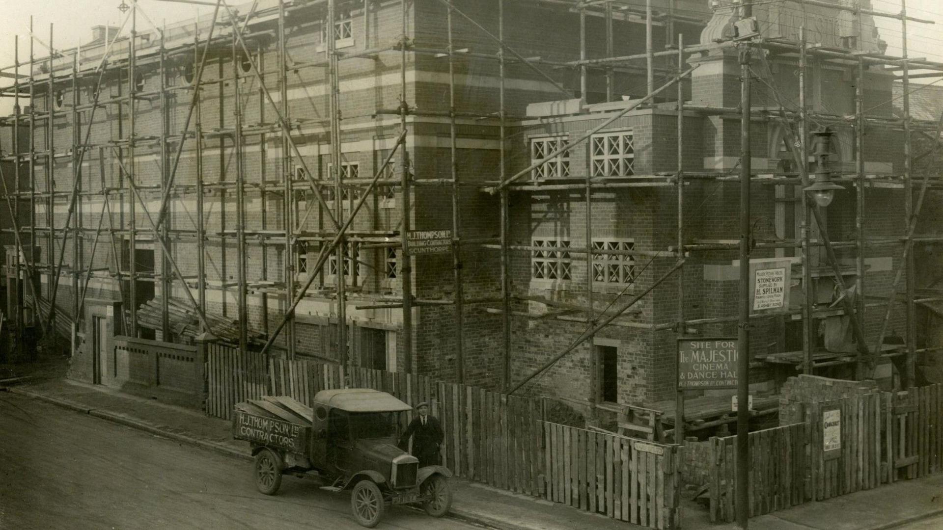 A black and white picture of a cinema building being built. There are large rows of scaffolding on the outside with a man standing next to an 19th century car.