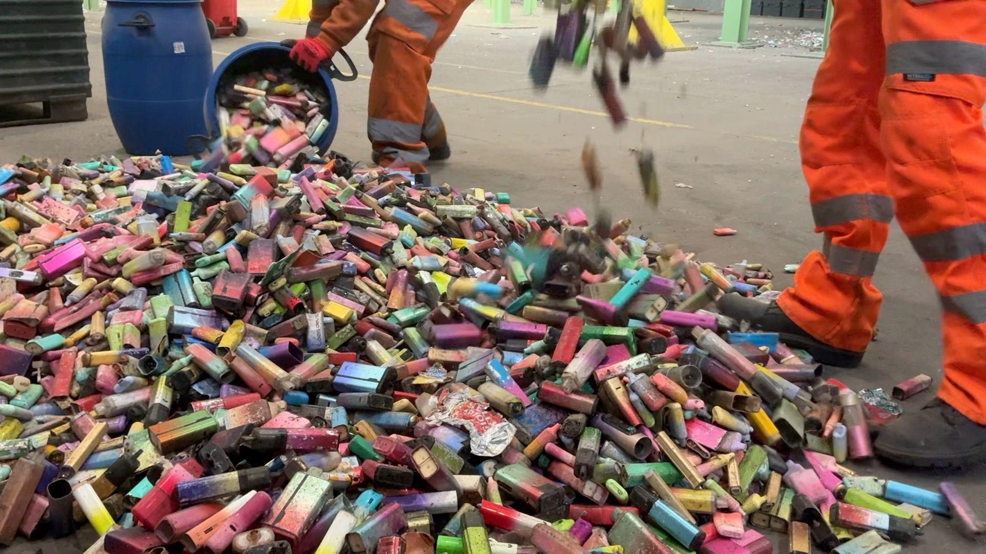A large pile of dirty used vapes on the concrete floor of a recycling facility. The vapes are in different shapes, sizes and colours. Two workers in orange overalls are adding to the pile from blue bins.