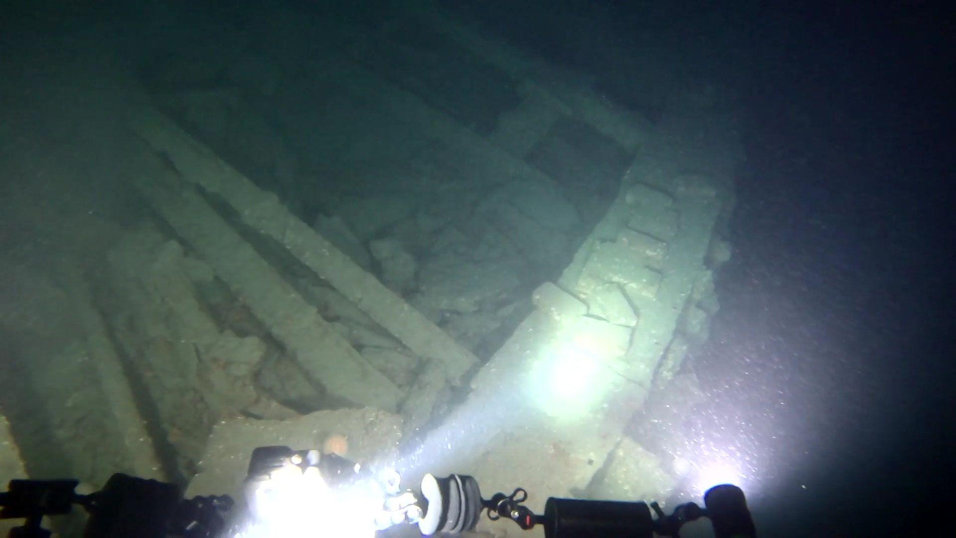 An underwater view of a wrecked ship. Two bright lights from a diver shine on to part of the wreck, which shows a small part of the hull and some joists.