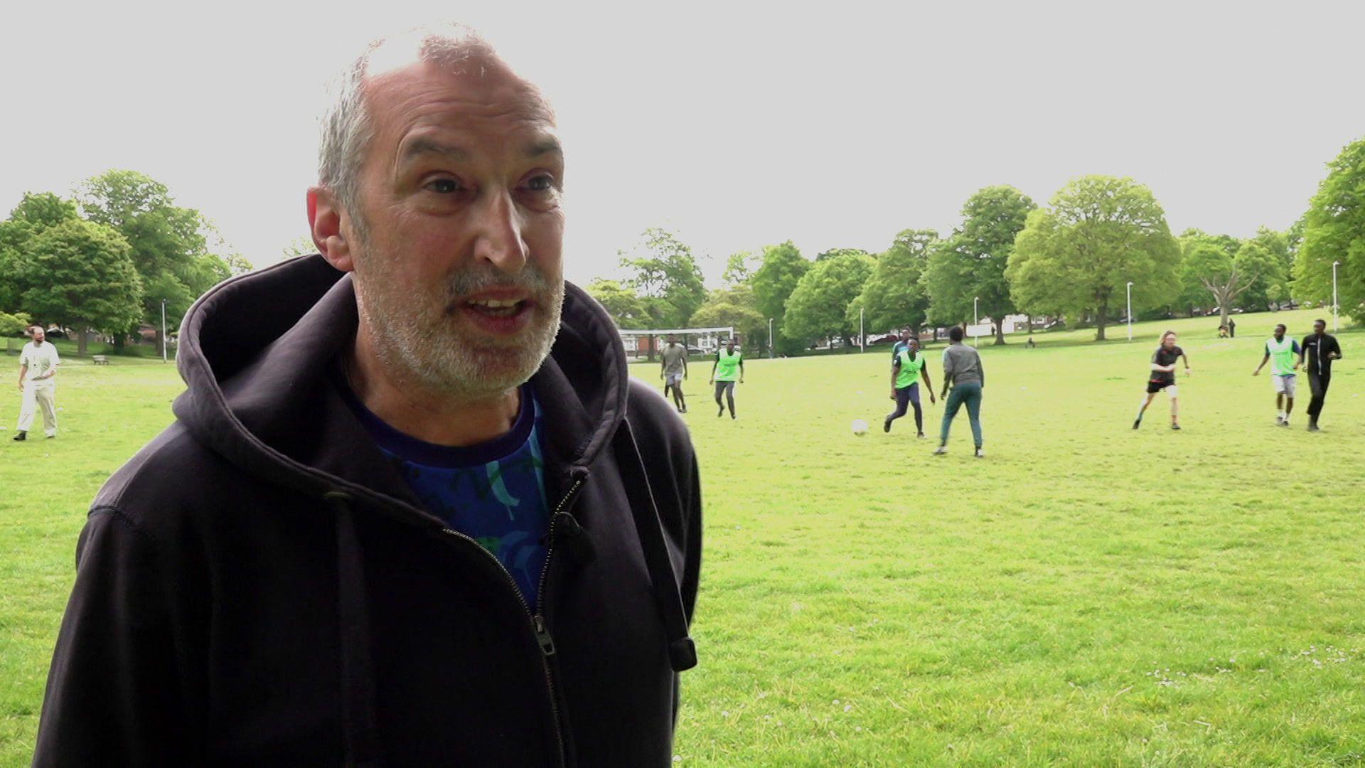 A man in a hoodie looks to the side of the frame while a game of football takes place in the grassy park behind him.