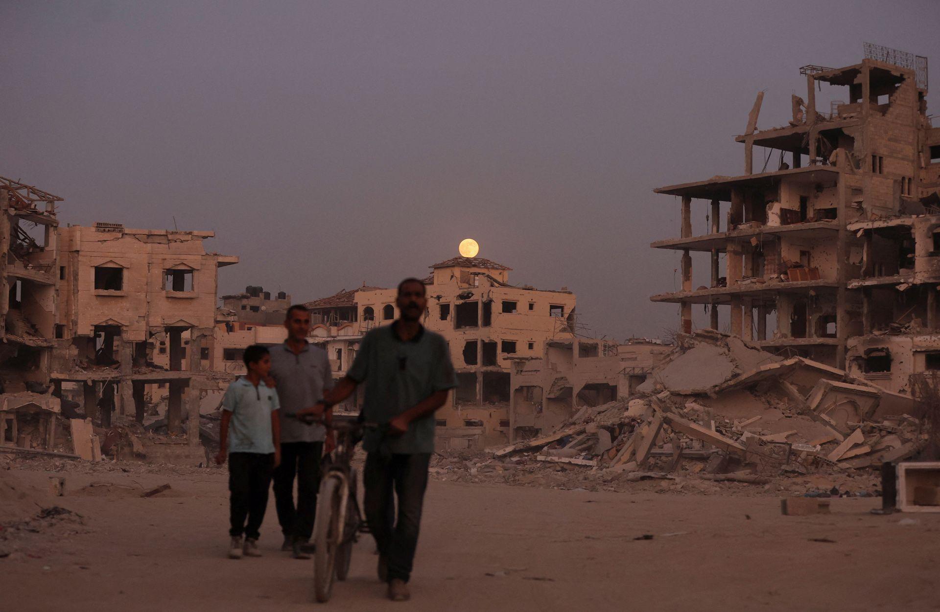 Silhouetted figures walk in the foreground while the Moon rises over a building in the background, with a destroyed building and rubble to the right, in Gaza City on Wednesday.