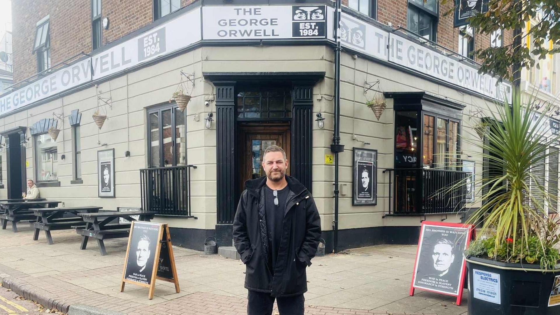 Daniel Davies outside 'The George Orwell'. He's smiling and has his hands in his pockets. He wears a black coat and has short hair and a short beard.