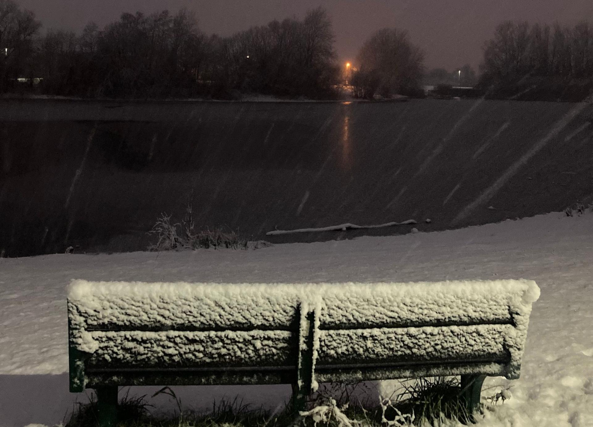 A snow covered bench next to a park lake. 