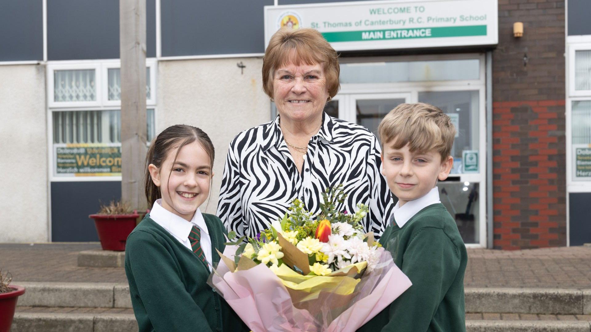 Mrs Hewlett stands outside the school with two children, they hold a large bouquet of flowers. She wears a zebra print shirt.