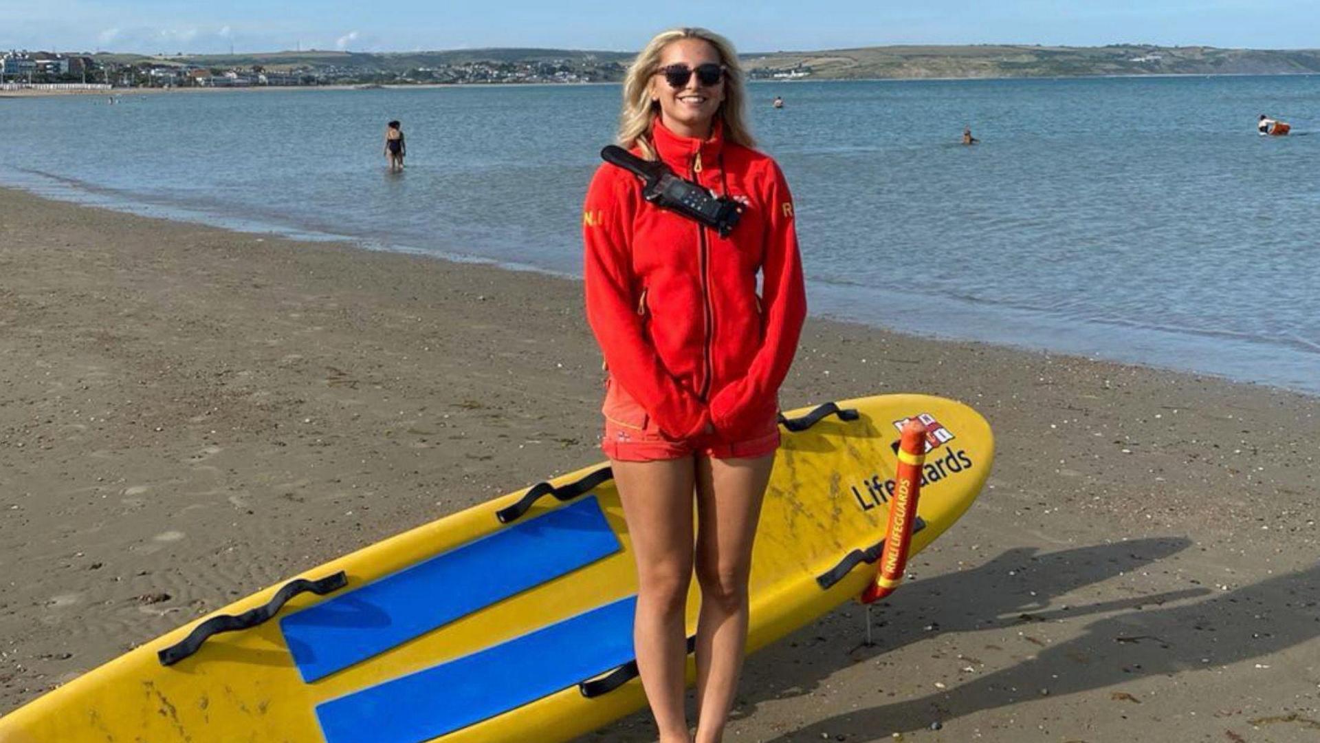 Molly is standing on the beach and smiling at the camera. She has her hands clasped together in front of her. She has long blonde hair which has been swept behind her shoulders and wears round dark framed sunglasses. She has a bright red zip up fleece on with the RNLI logo on the right side of her arm. She's got a black walkie talkie strapped around her chest. In the background is the sandy beach, sea and cliffs in the distance. There are a couple of people in the sea behind her.