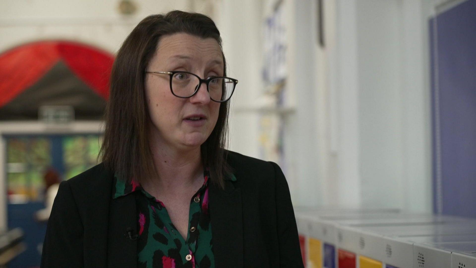 A woman with dark, long hair, wearing glasses, sits in a classroom speaking to the right of the camera