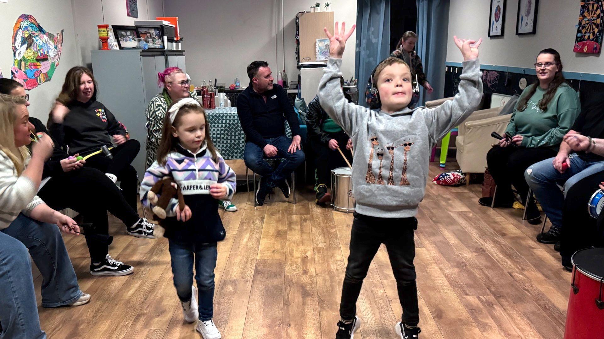 A young girl on the left and a young boy on the right at the autism group. The girl is holding a toy monkey and the boy is wearing a grey jumper with giraffes on and raising his arms in the air. Around the edge of the room adults watch on.