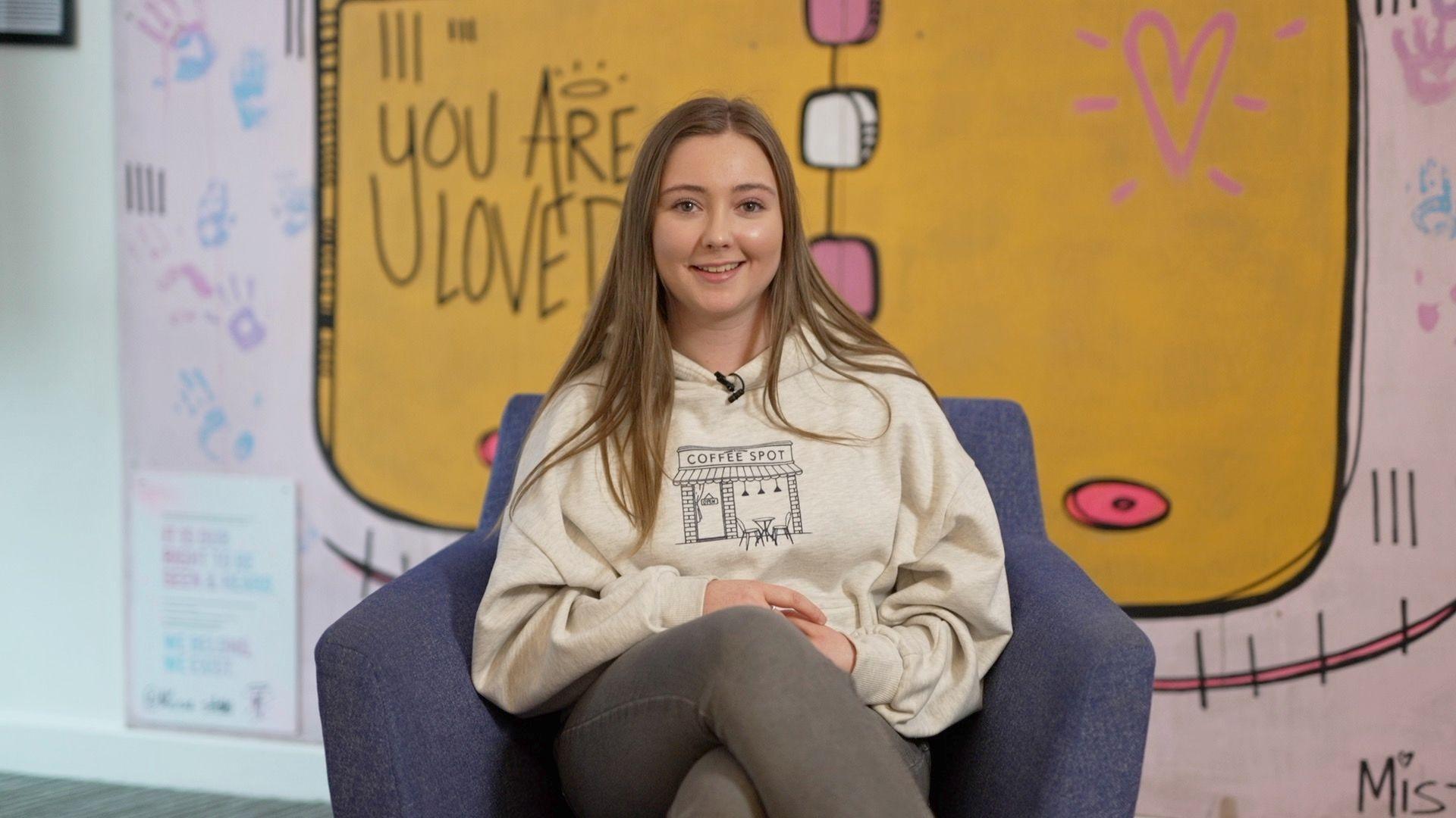 Hannah Clubley sits in a blue tub chair in front of a yellow sign that says 'you are loved'. She is wearing a cream hoodie and grey jeans Her blonde hair is over her shoulders. 