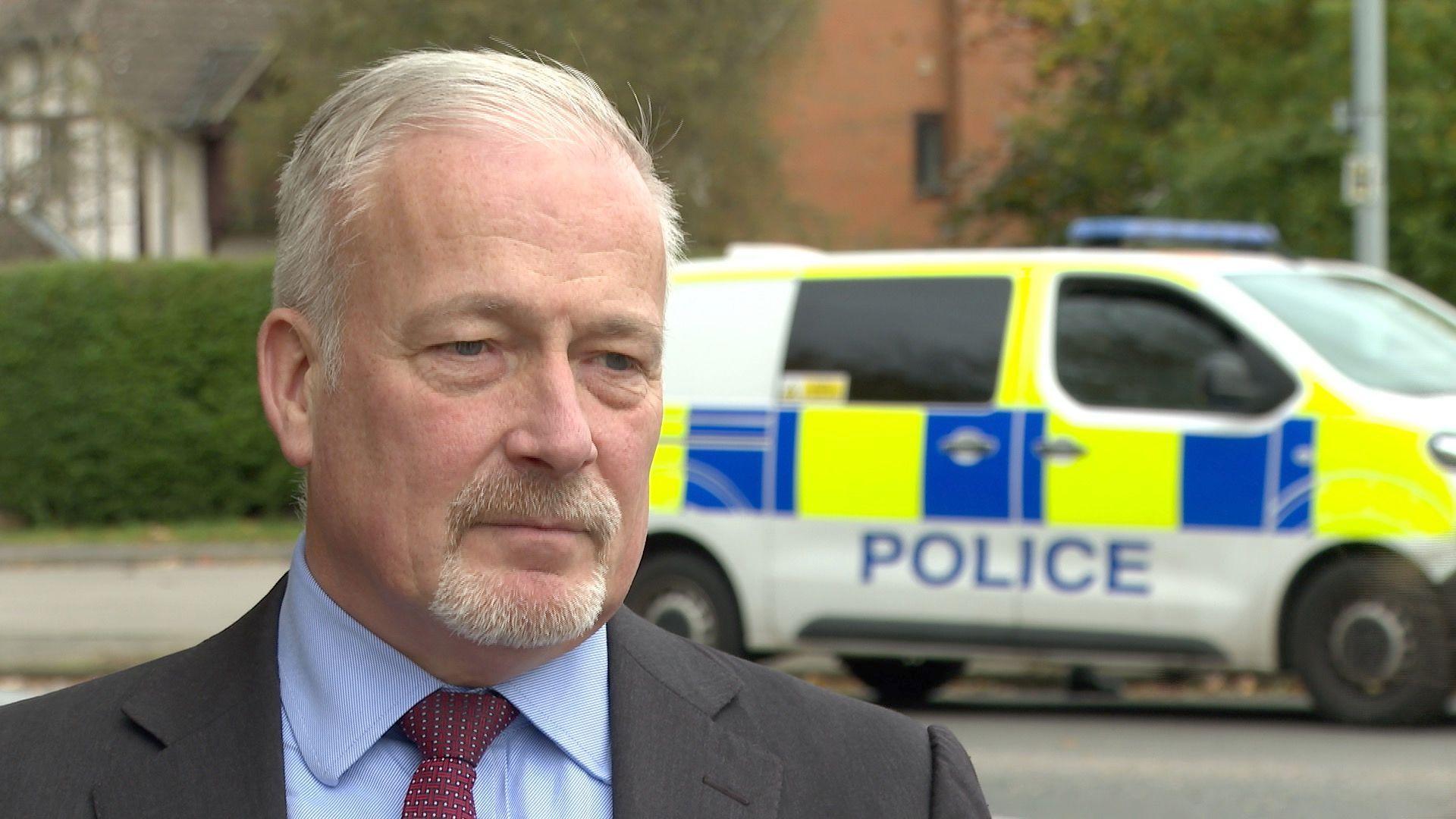 Richard Fuller, who has short white hair and a goatee, is standing in a street near a police van. He is wearing a charcoal suit, a blue shirt, and blue tie with white dots.