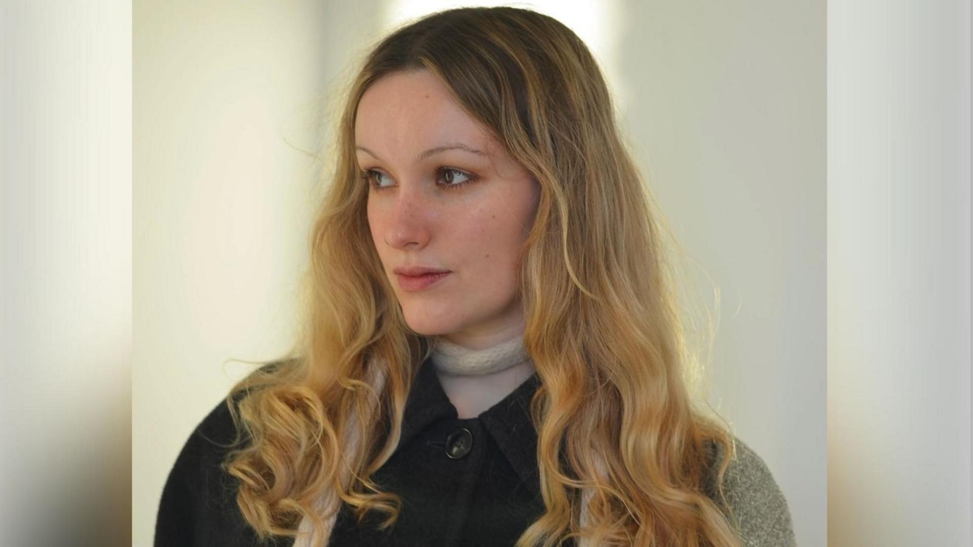 Aimée MacDonald is pictured looking to the left of the image with a neutral expression. She has long blonde hair and wears a black coat with a thin white scarf round her neck.