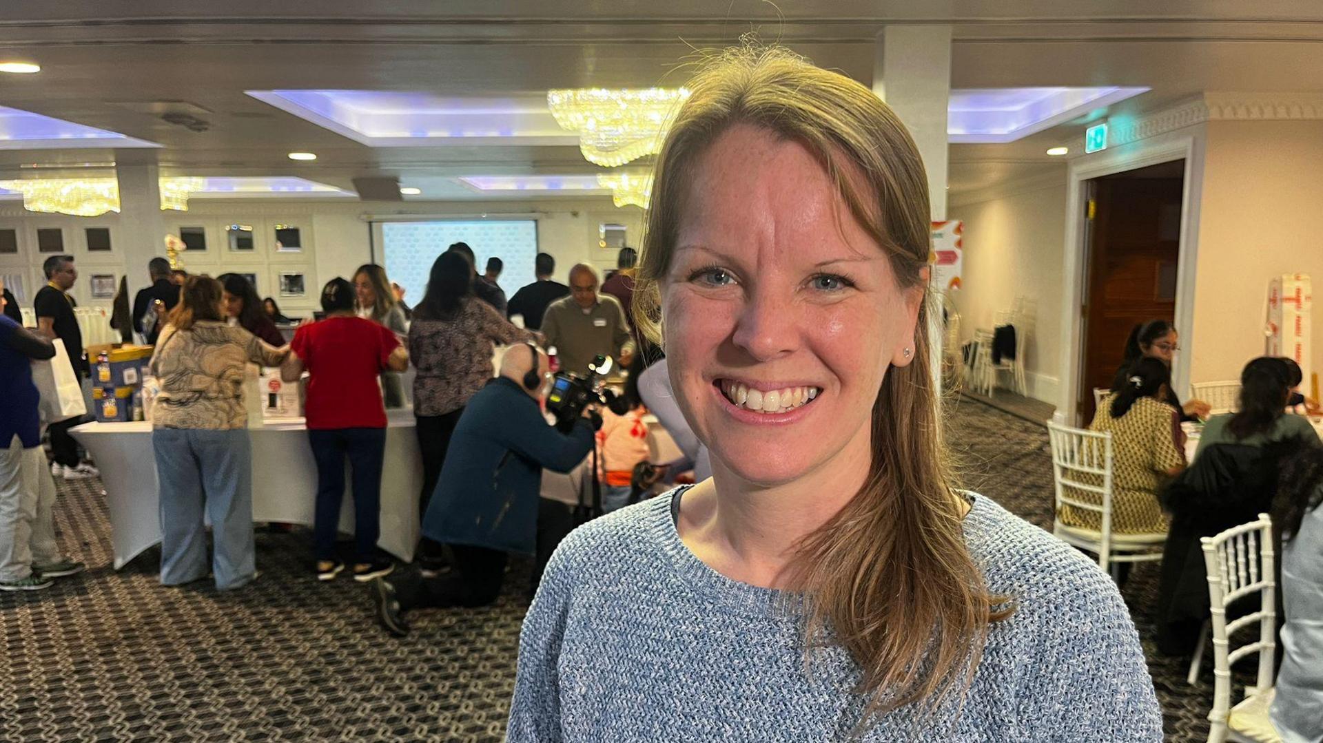 Emma Rapley is standing in the middle of a room were volunteers are putting together the packages. She is wearing a grey knitted jumper and is smiling at the camera.