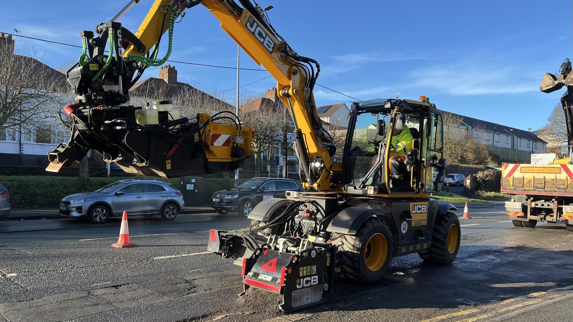 A yellow and black JCB machine on the edge of a suburban A road with parked cars and houses in the background. The area in which it is working is separated off by traffic cones. The machine is planing the tarmac with a cutter on the front. the driver, wearing a yellow hi-vis jacket is visible siting in the cab. 