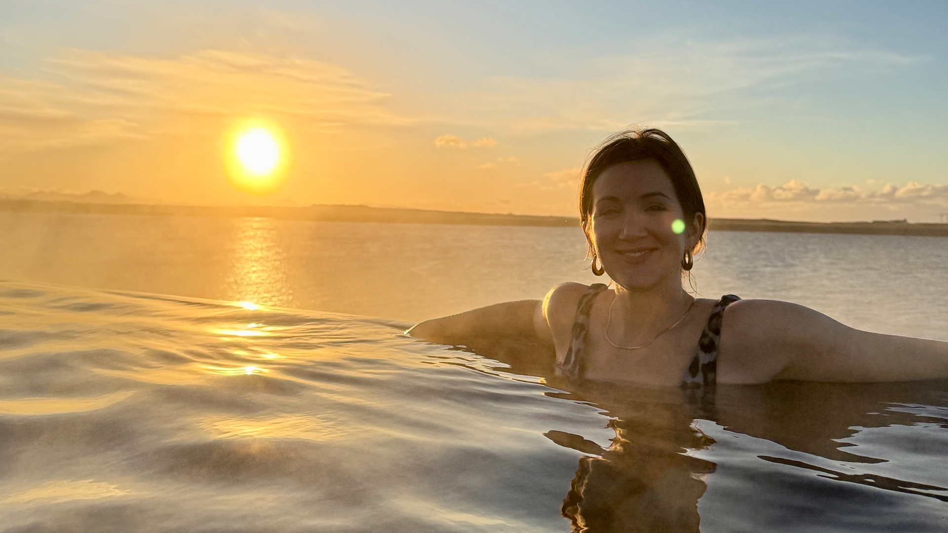 Monica Stott sits in a pool at the Sky Lagoon, in Iceland. The sun is setting on the left of her. 