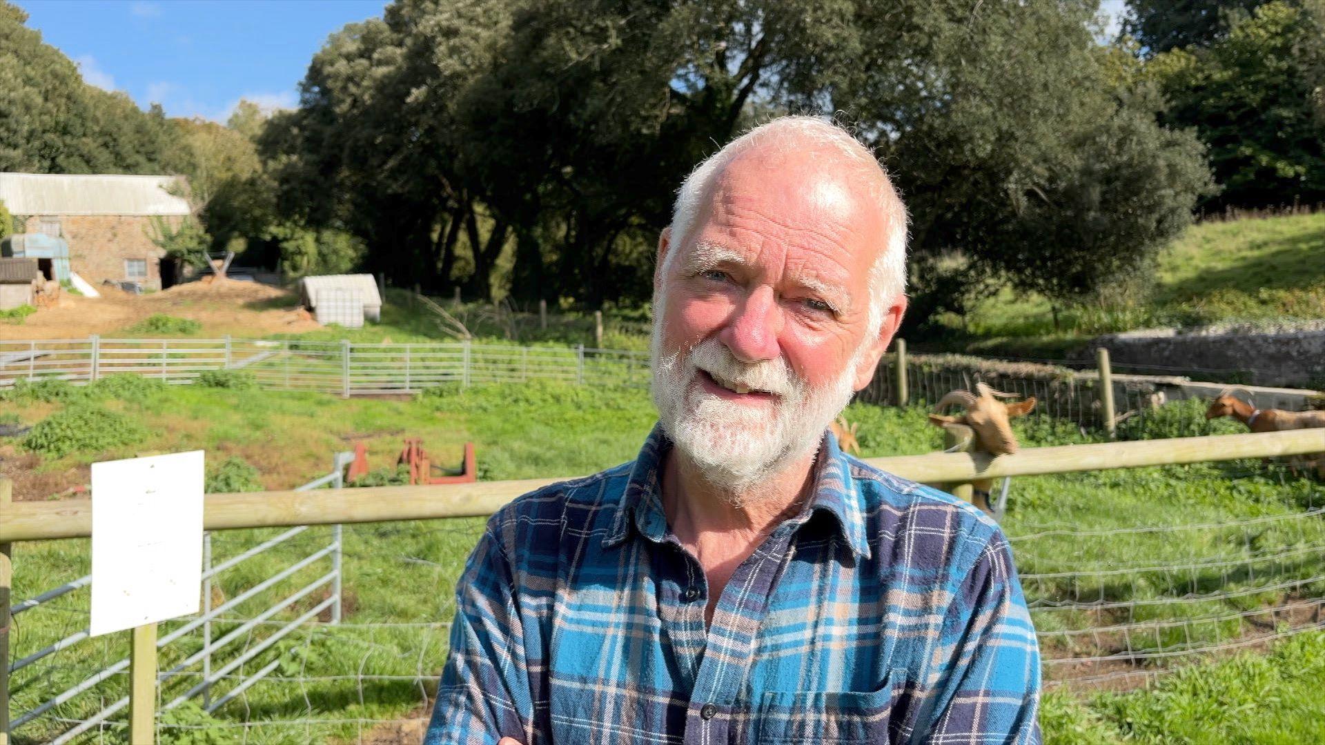 A man with a white beard, wearing a blue checked shirt stands near Golden Guernsey Goats on his farm.