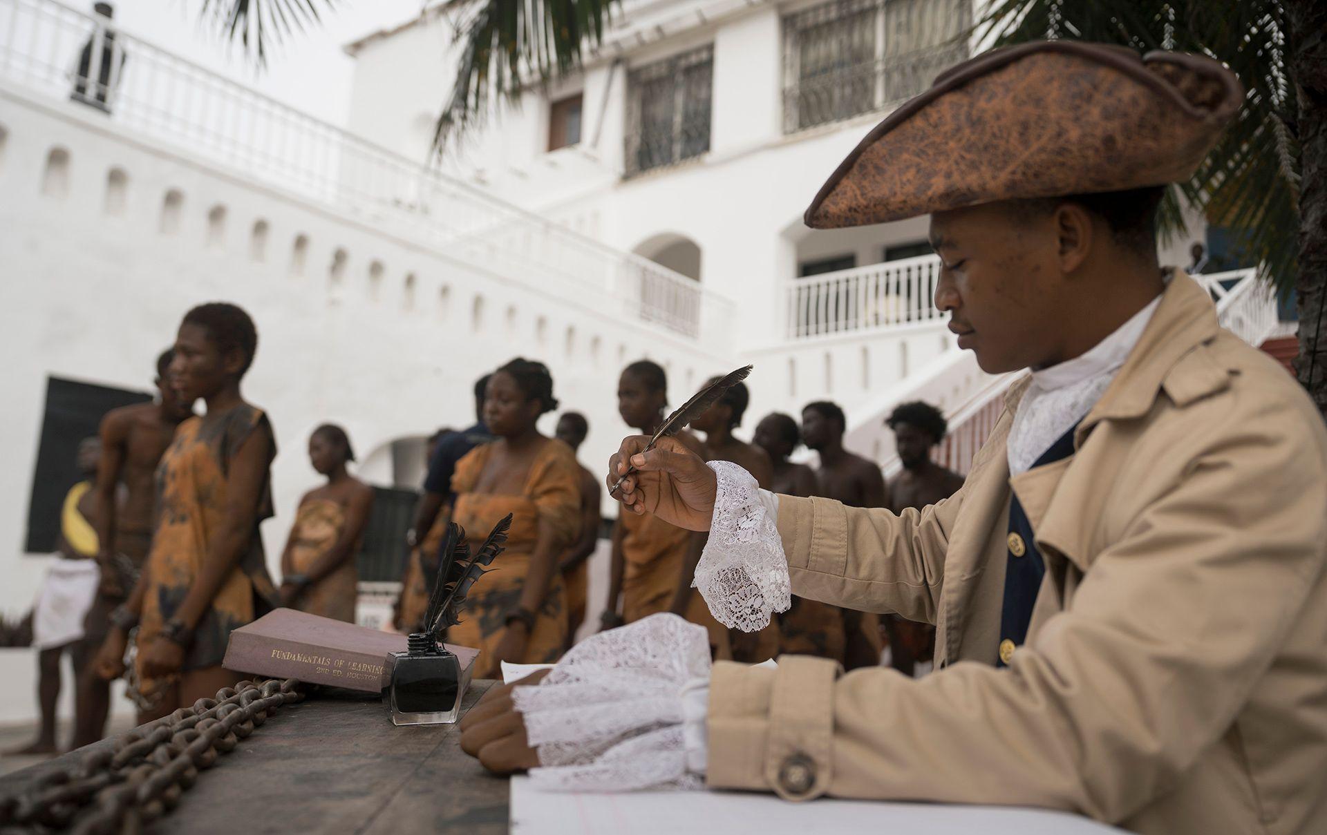A person in the foreground wears pre- Victorian-era clothing and signs a document with a quil while wearing a hat, as people dressed in scantily clad slave robes walk through the street in chains in the background, during a re-enactment of the trans-Atlantic slave trade in the courtyard of Christiansborg Castle in Accra, Ghana on 19 June.