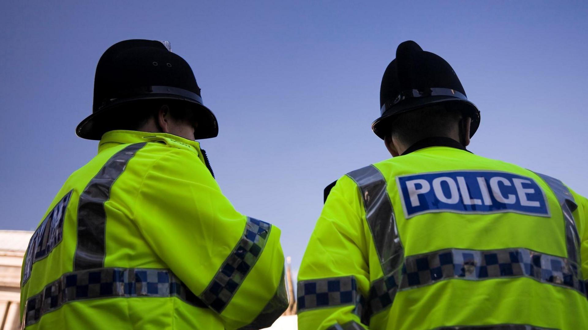 The back of two police officers wearing green jackets with "police" written on in white writing against a blue background. 