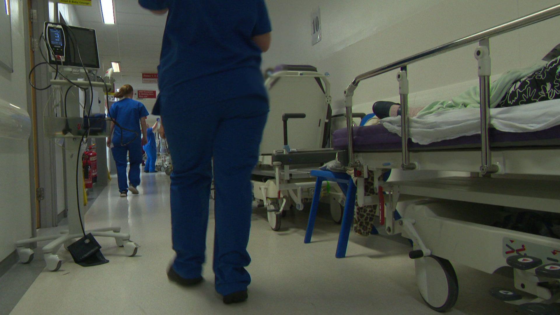 A nurse in blue scrubs walks down a hospital corridor past several patients lying in hospital beds.